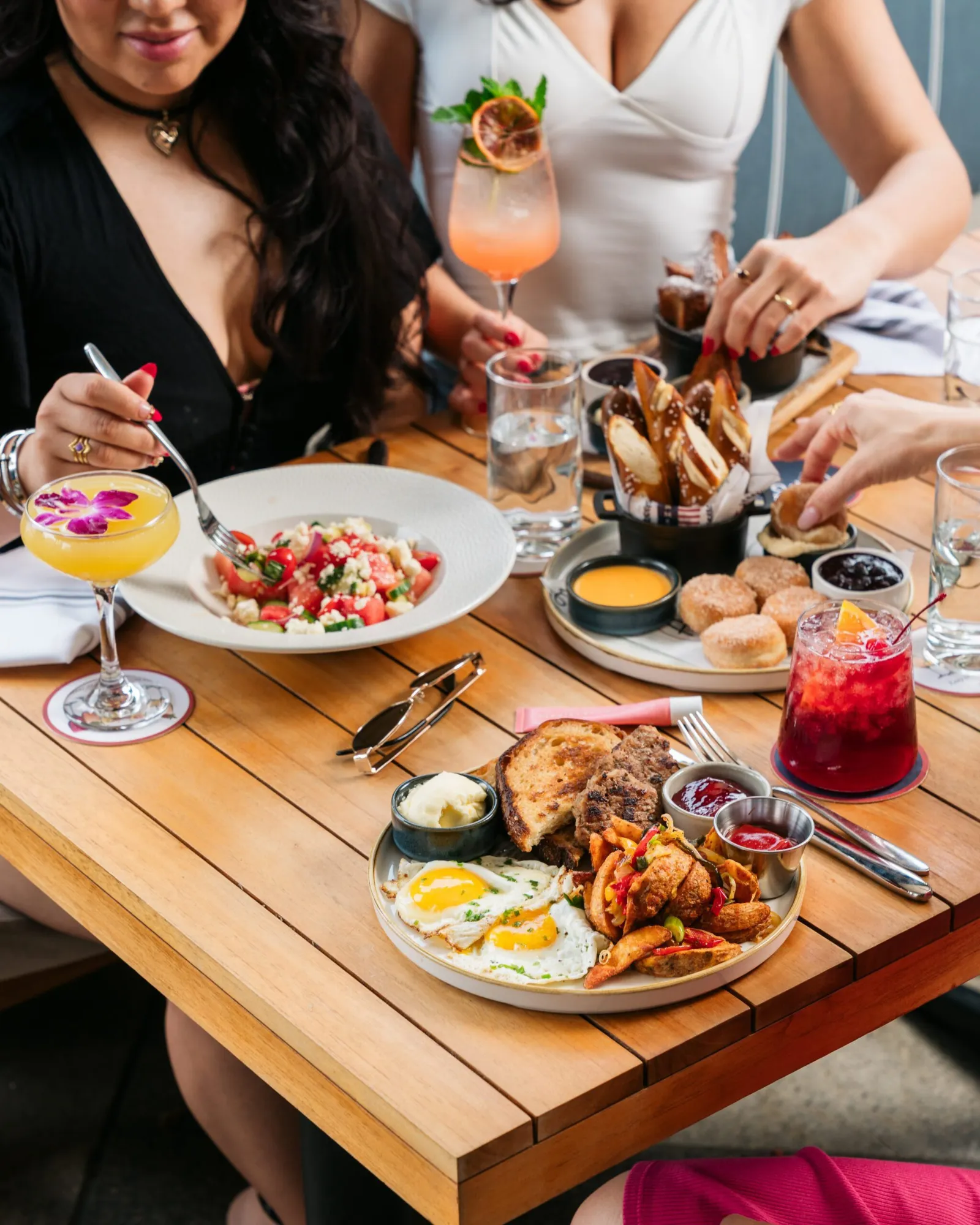 Three women sit at a wooden table enjoying brunch dishes and colorful drinks, including fried chicken with eggs, a salad, toast with spreads, and cocktails. Hands reach for food, creating a lively, so