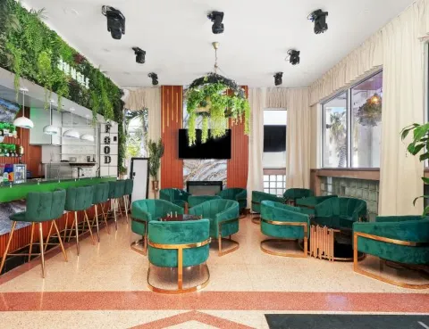 An indoor bar lounge with green velvet seating, a counter, pendant lights, and cascading plants filling the space.