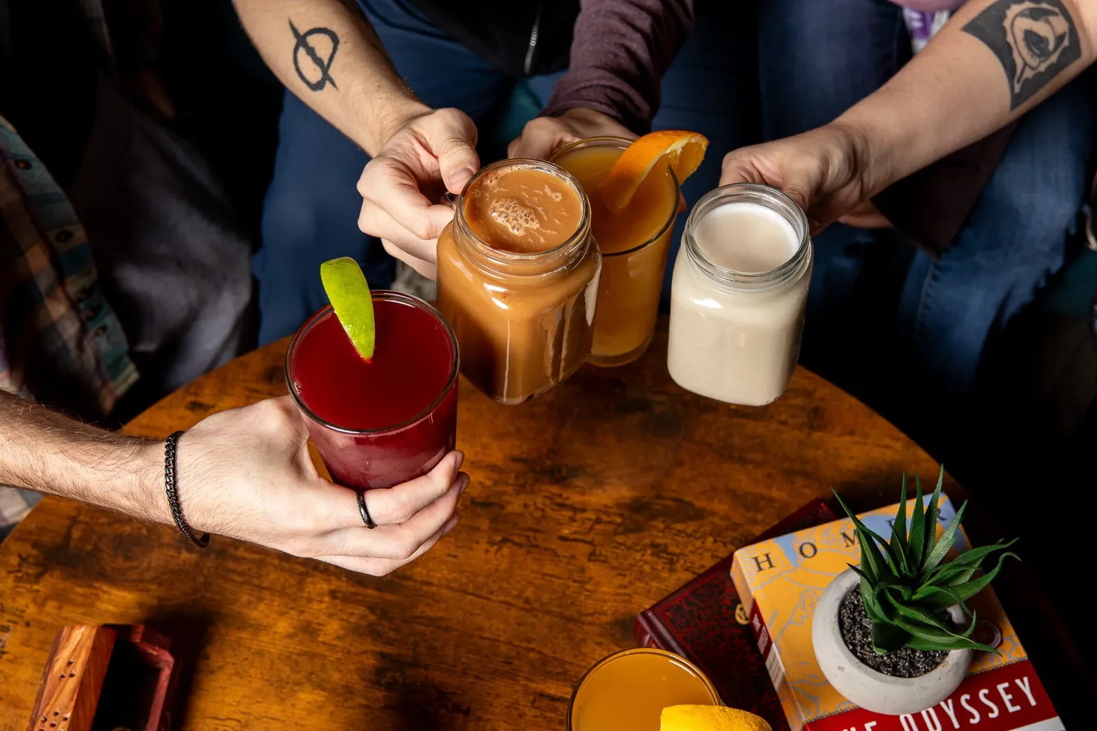Four people toasting with colorful beverages, including a red drink with lime, an orange mocktail with an orange slice, a frothy brown drink with a cinnamon stick, and a white milkshake, on a wooden t