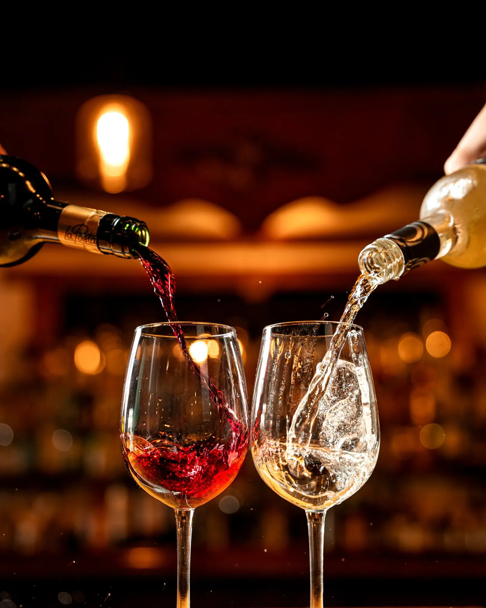 Two wine glasses, one filled with red wine and the other with clear white wine, being poured from bottles with blurred warm lights in the background.