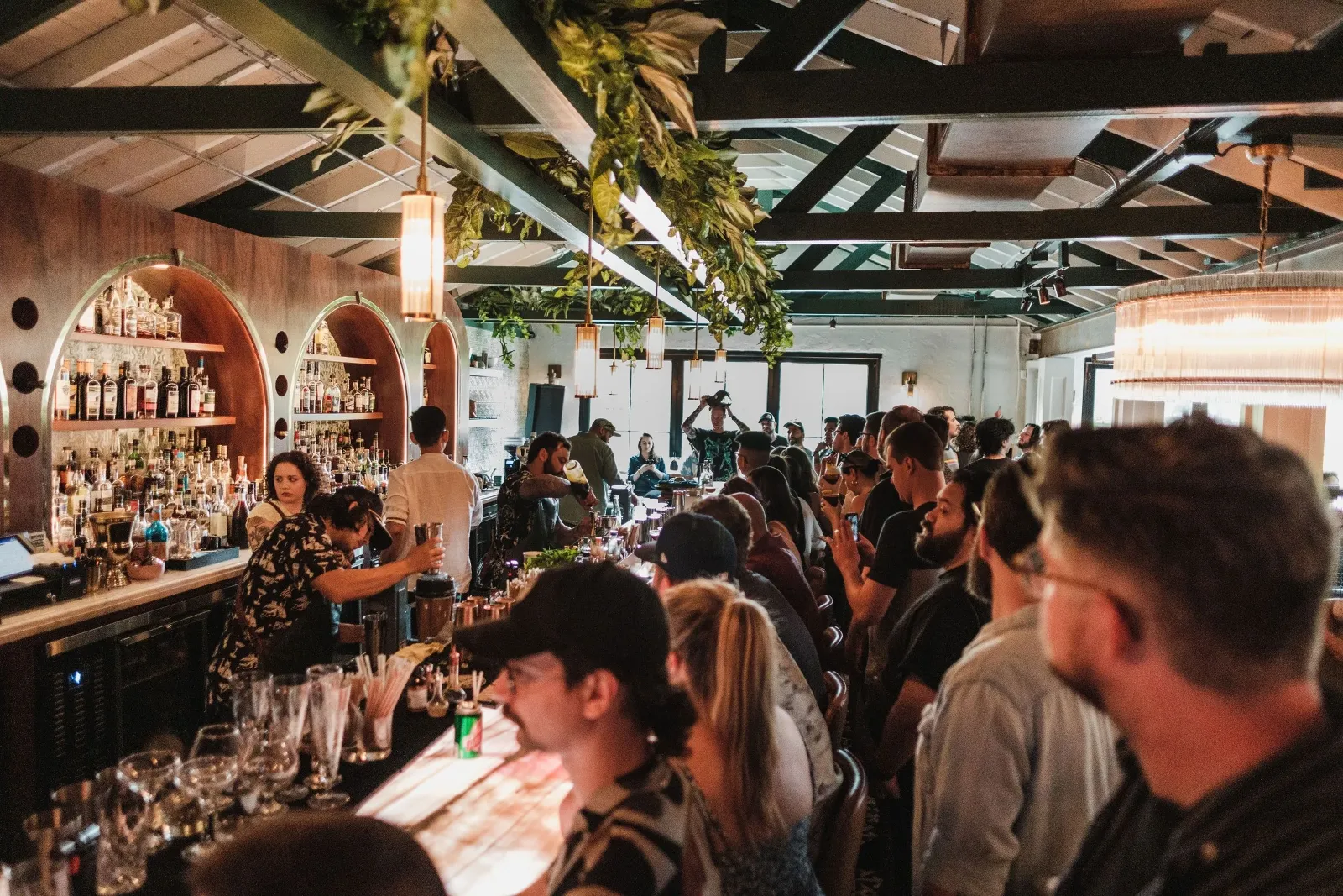 Crowded bar with people socializing, barista preparing drinks, shelves of liquor bottles, hanging plants, and warm lighting.
