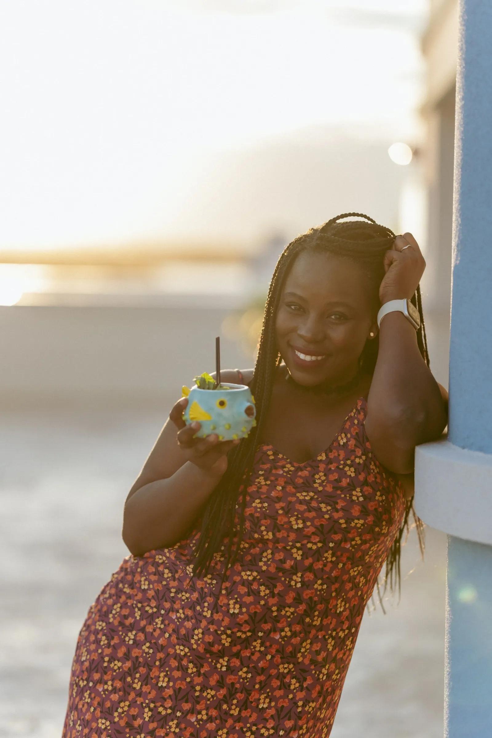 A woman with braided hair wearing a floral dress, standing outdoors at sunset, holding a tropical drink mug, and smiling.