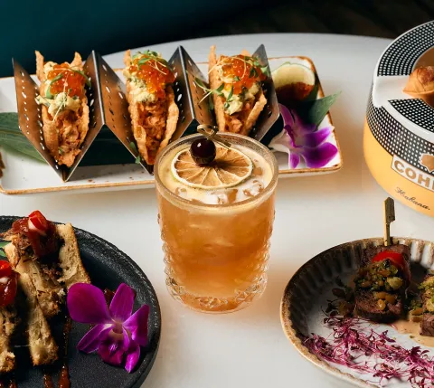 Iced citrus cocktail with lemon slice and cherry, surrounded by tapas and fried bites on a bright table.
