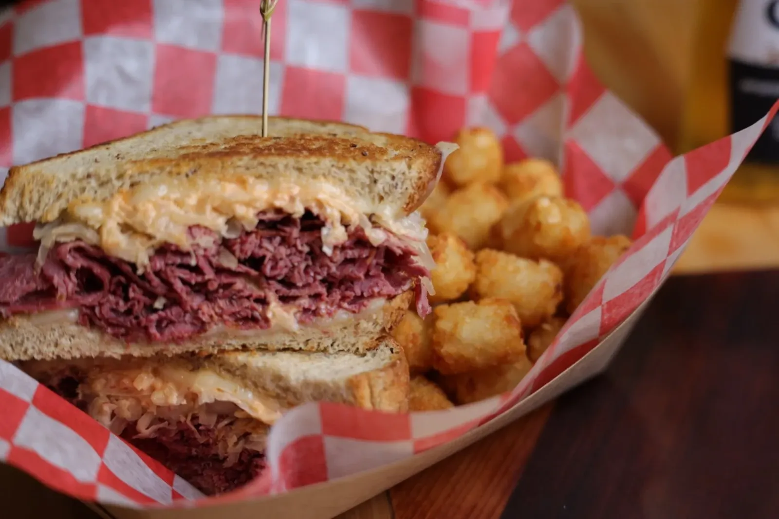 A sliced Reuben sandwich with corned beef, sauerkraut, and melted cheese on toasted bread, served with tater tots in red and white checkered paper.