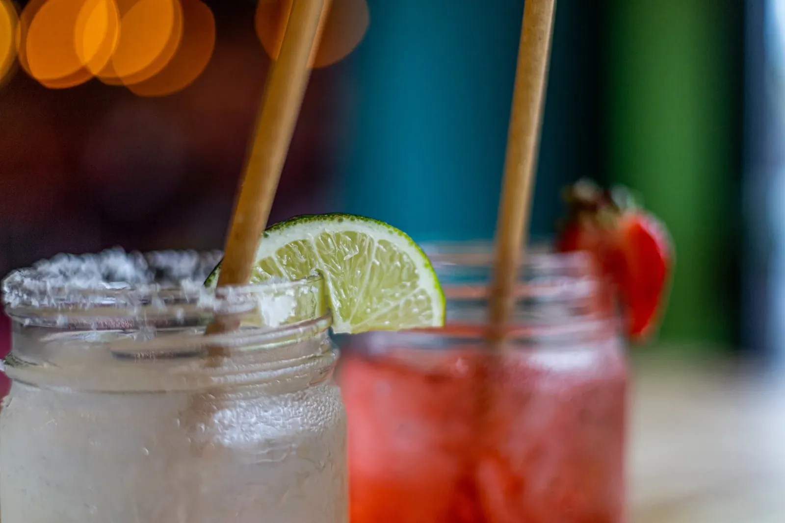 Two mason jars with fruit drinks, one with a lime wedge and the other with a strawberry, both with brown straws, set against a colorful blurred background.