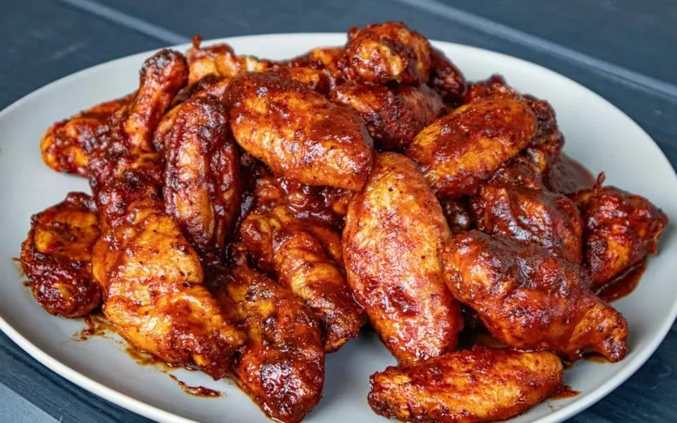 A pile of crispy, red glazed chicken wings on a white plate against a dark, wooden background.