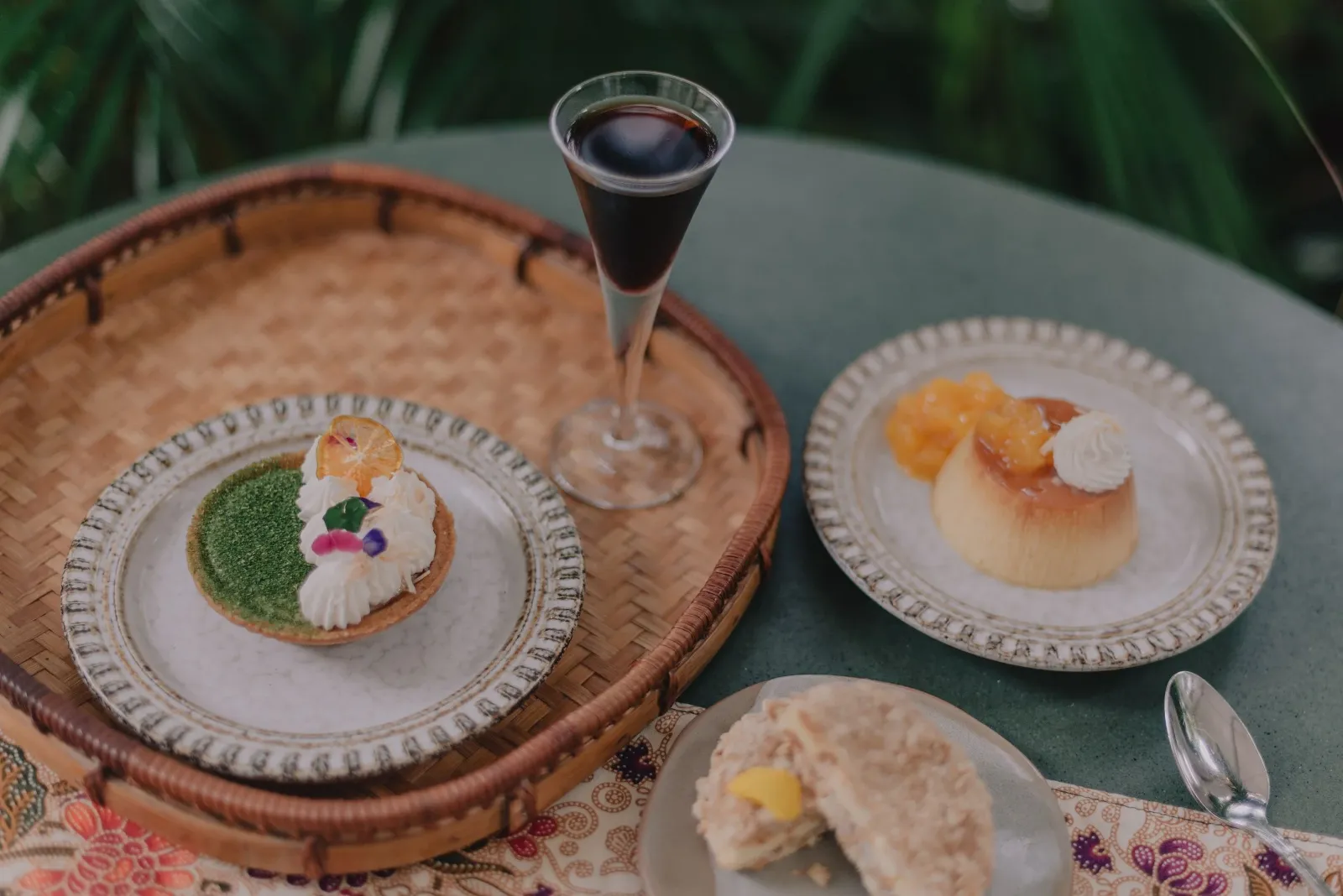 Assorted desserts and a glass of red wine on a table. A green and white pastry on a plate, a custard dessert with fruit topping, and a pie slice with fruit filling.