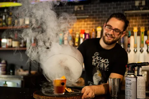 A barista wearing glasses and a black t-shirt smiling as he prepares a cocktail with dry ice, creating a fog effect in a bar setting.
