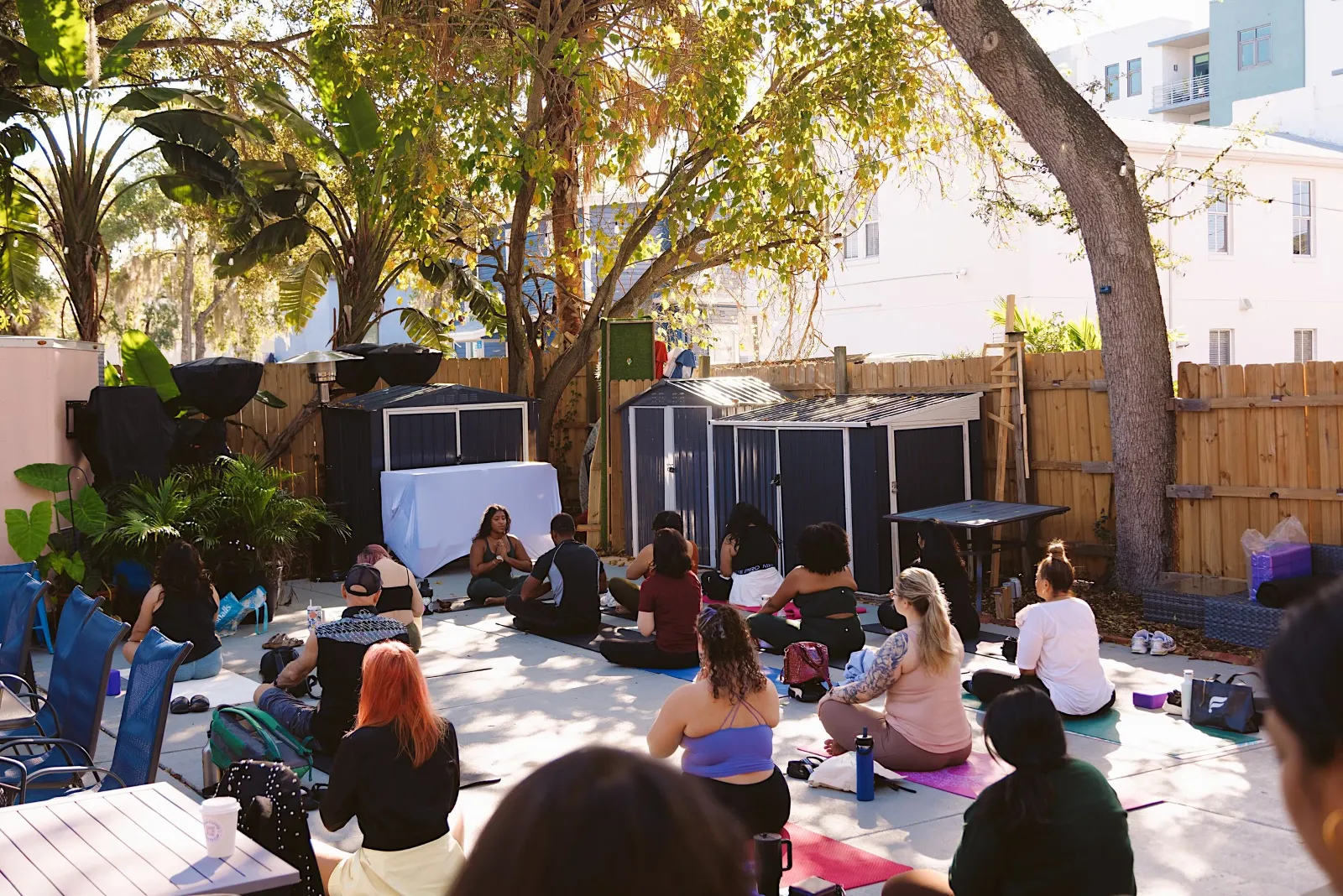 People participating in a yoga or meditation class outdoors in a backyard with trees and a wooden fence.