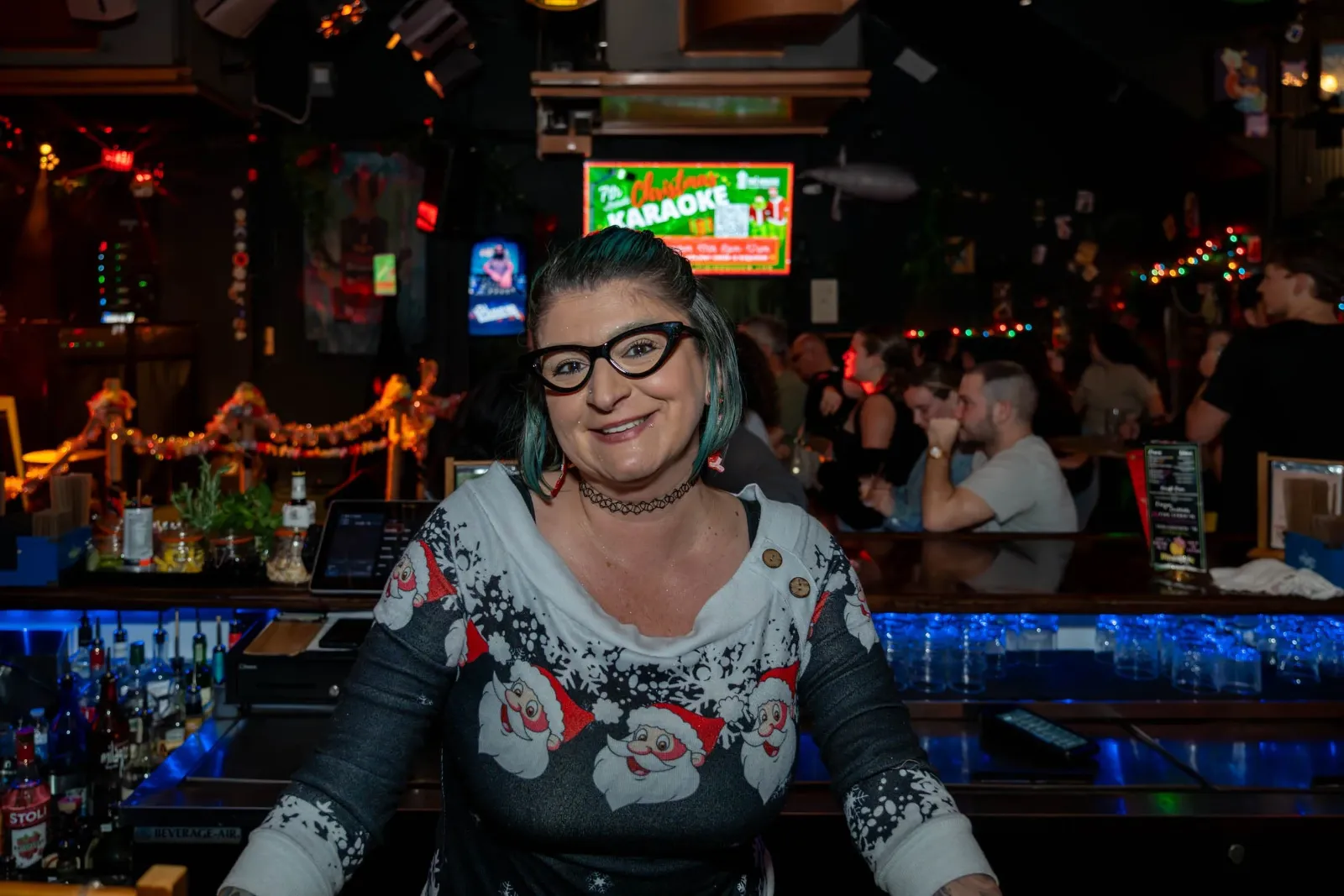 A woman with glasses, black choker, and Christmas sweater smiles behind a bar in a lively, decorated pub with patrons in the background.