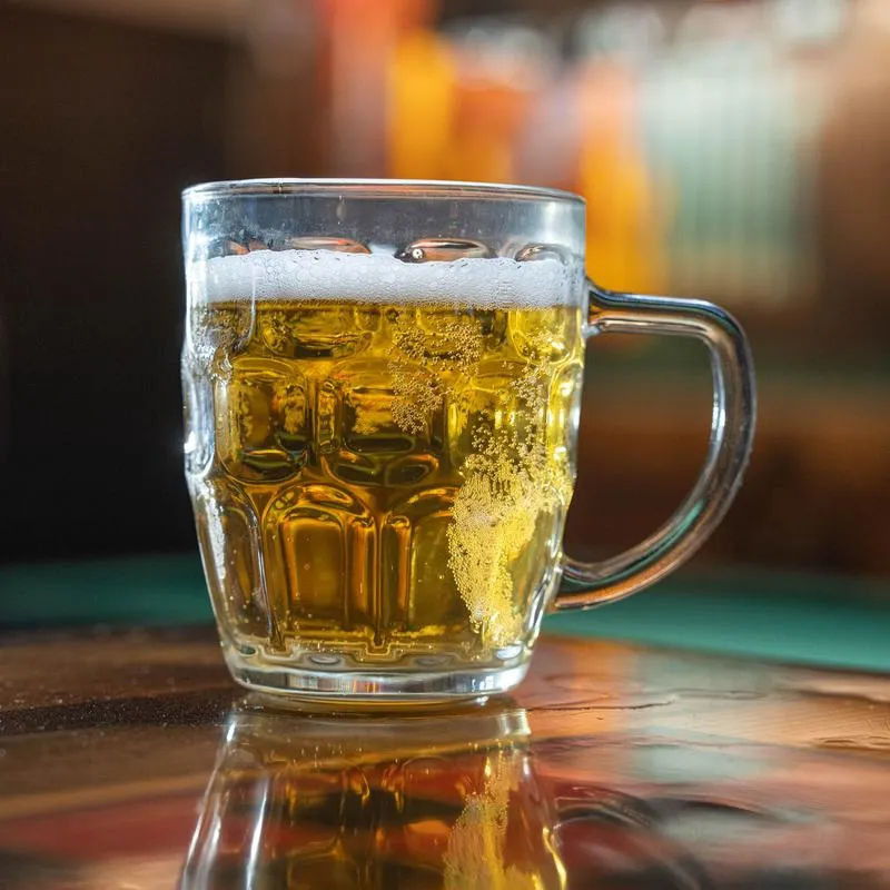 Golden beer in a clear glass mug with foam on a wooden bar counter.