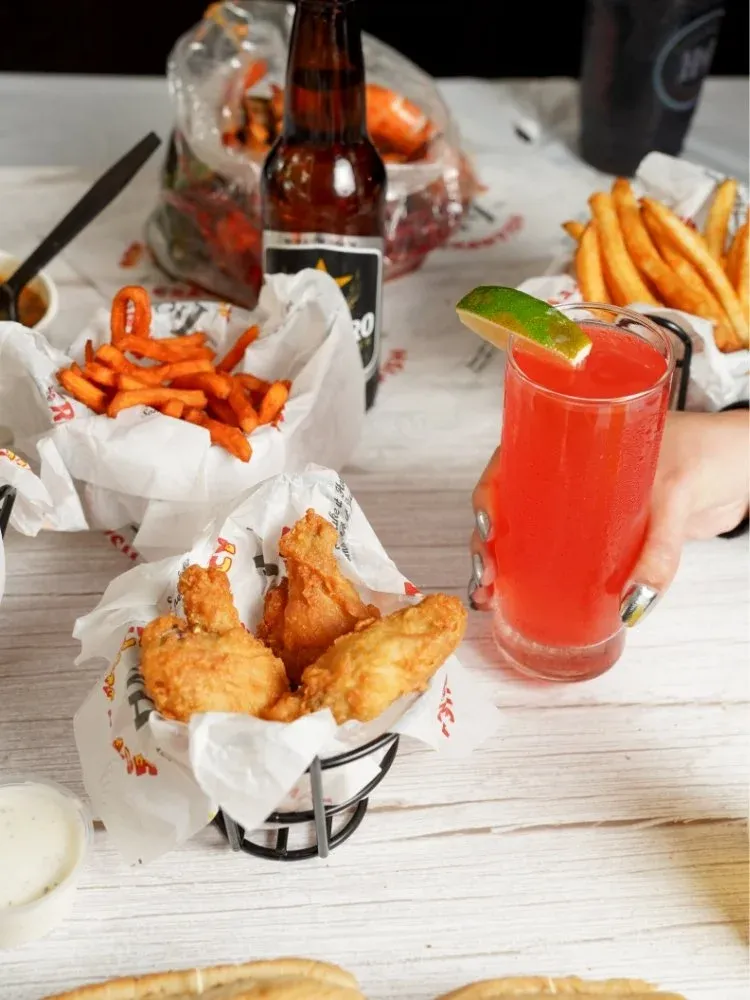 Fried chicken tenders, curly fries, and a red cocktail with a lime wedge on a white wooden table at a restaurant.