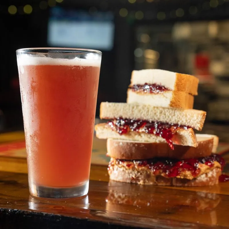 Glass of fruit beer and peanut butter and jelly sandwiches on a wooden counter.