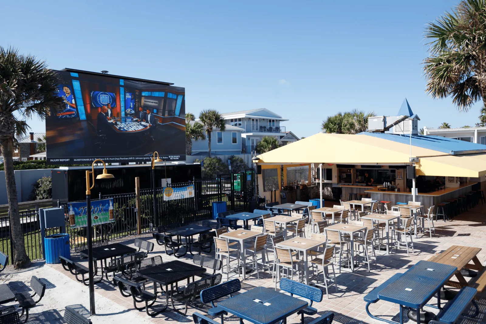 Outdoor bar and seating area with tall tables and chairs, large yellow umbrellas, and a large TV screen showing a sports talk show, surrounded by palm trees and modern buildings in the background.
