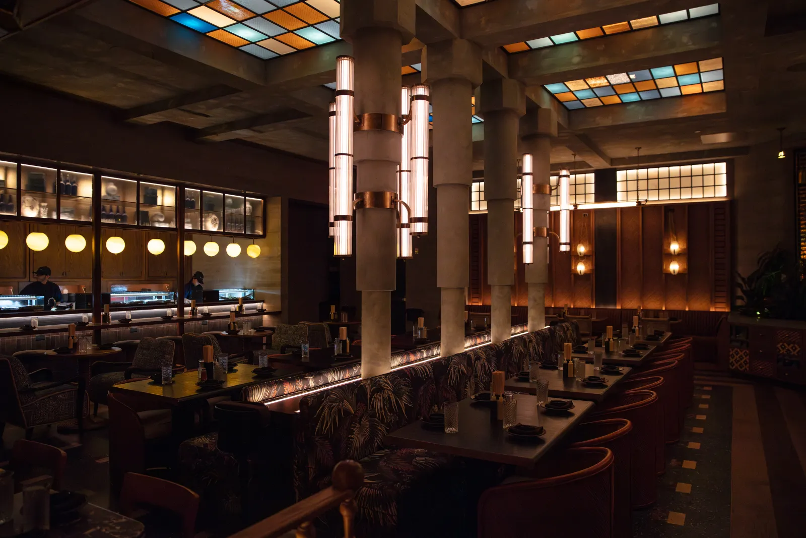 Interior of a restaurant with modern design. There are tables with dark napkins, and chairs with patterned fabric. The ceiling has colorful stained glass panels, and hanging cylindrical light fixtures