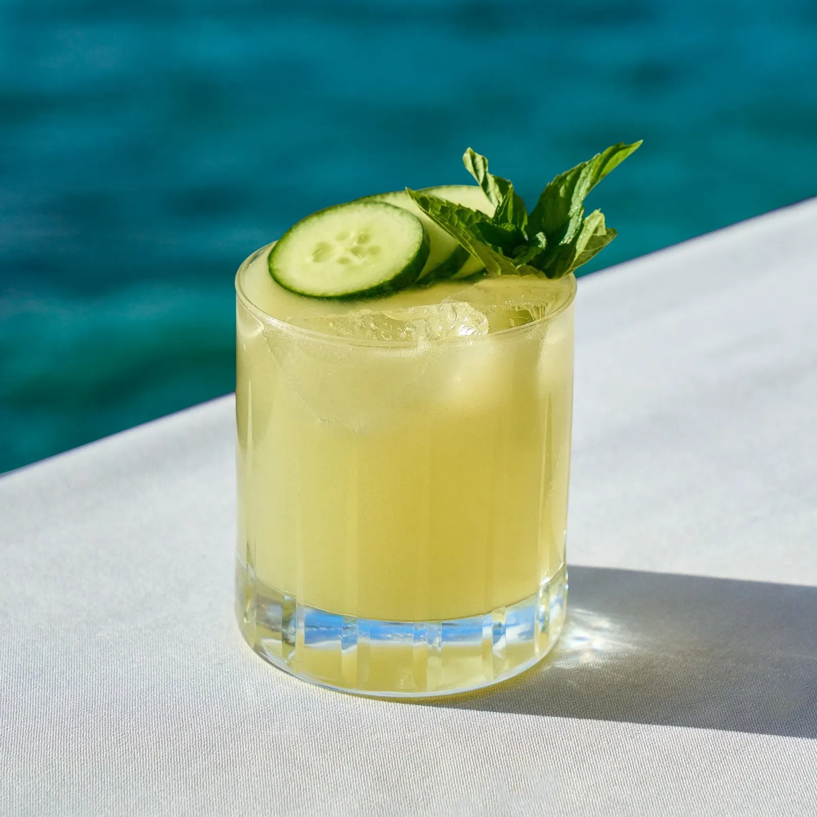 Cucumber cocktail garnished with cucumber slice and mint in a glass with ice, placed on a white surface with a blurred blue background.