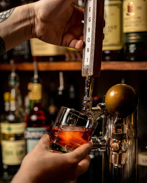 A person holding a glass under a tap pouring a pinkish drink at a bar, with bottles on shelves in the background.