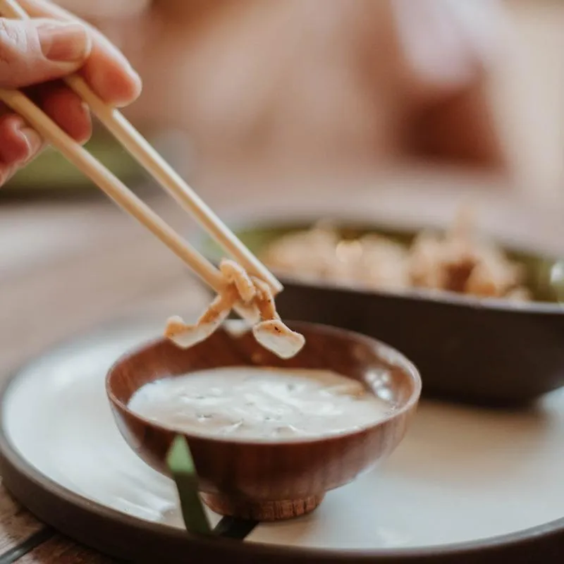 A person using chopsticks to dip food into a creamy white sauce.