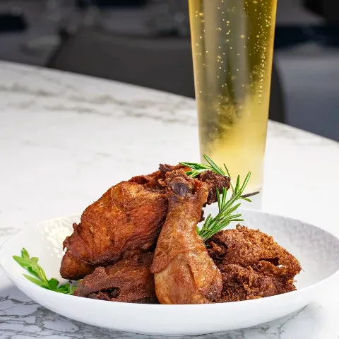 Golden, crispy C & C 48 Hour Chicken served on a white plate with fresh rosemary garnish, paired with a cold draft beer on a marble bar at Catch & Cut Seafood & Steak. The softly blurred dining room c