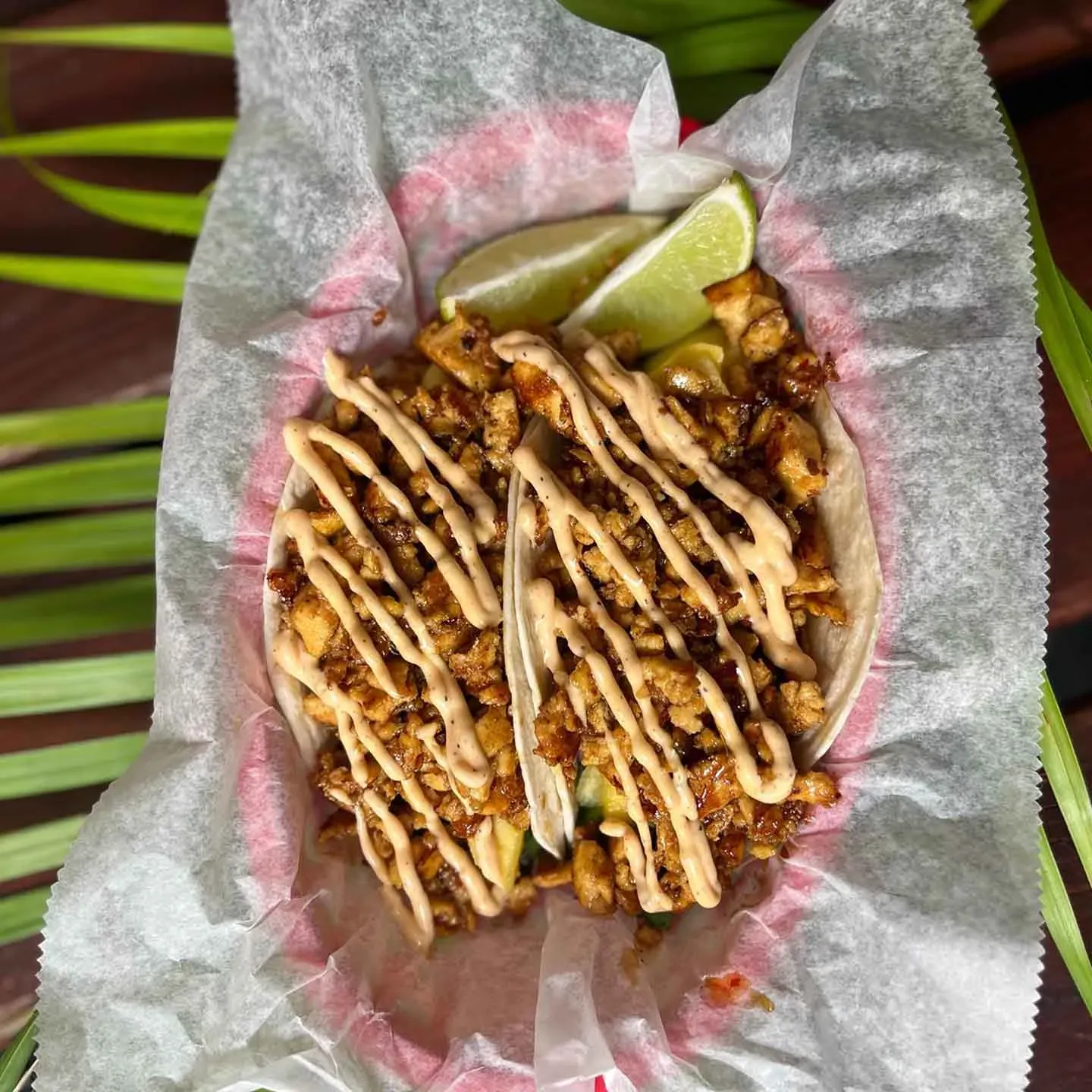 Two tacos filled with seasoned meat, topped with creamy sauce, served with lime slices in a paper-lined basket.