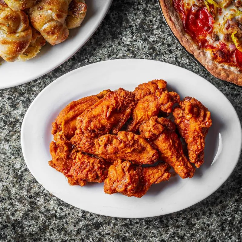 Buffalo wings served on a white plate, accompanied by a pizza and garlic knots.