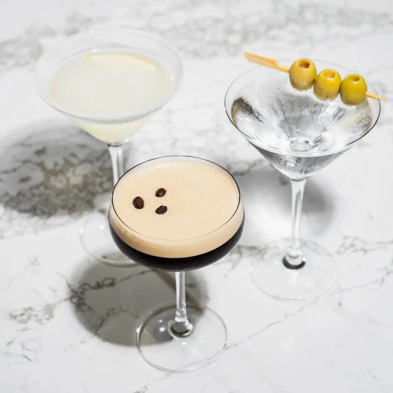 Three cocktails in martini glasses on a white marble surface. One drink is deep brown, the second is light, topped with two coffee beans, and the third is pale white with three green olives on a skewe