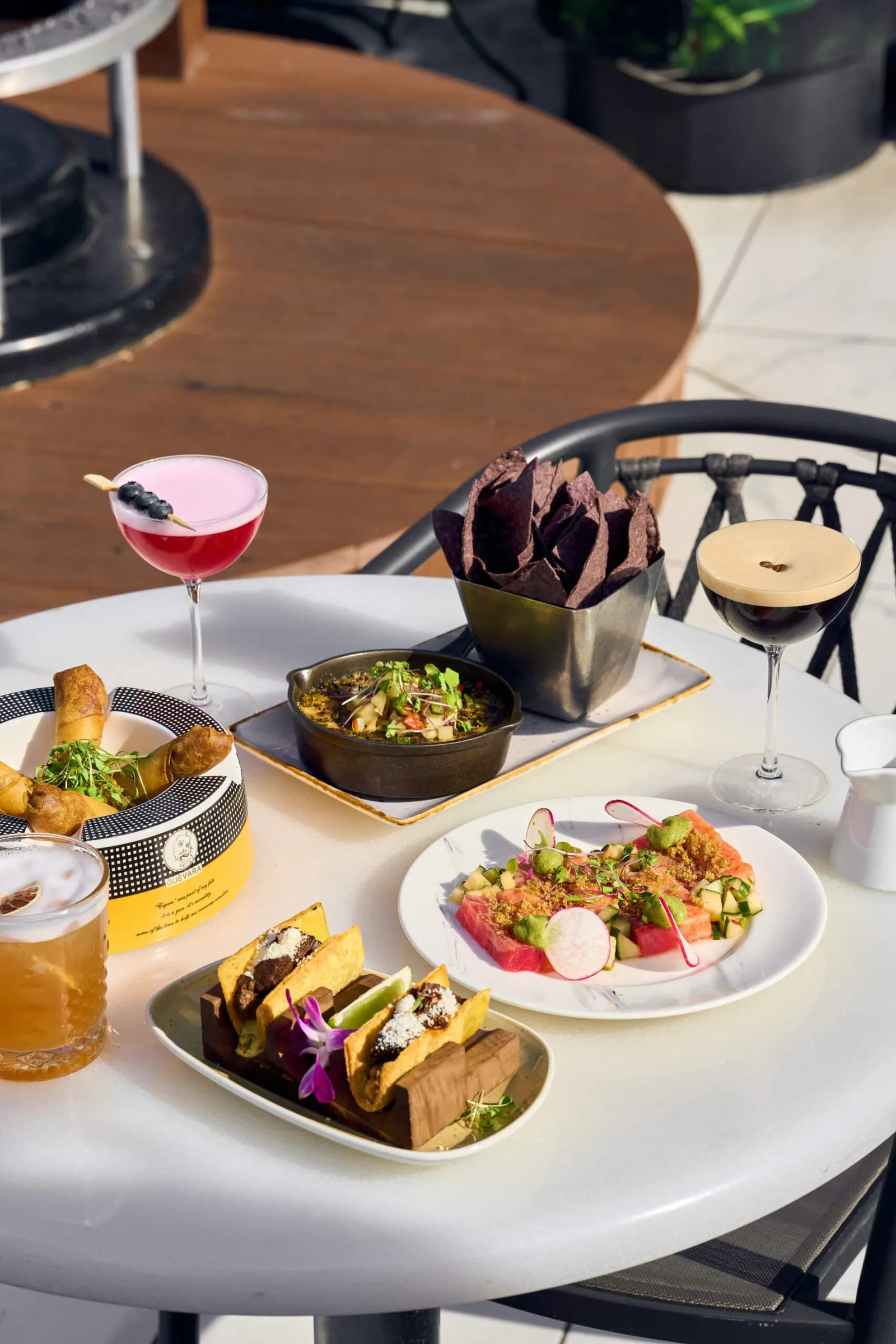 Outdoor table with a variety of tapas-style dishes and two cocktails, topped with chocolate chips and herbs, on white tableware.