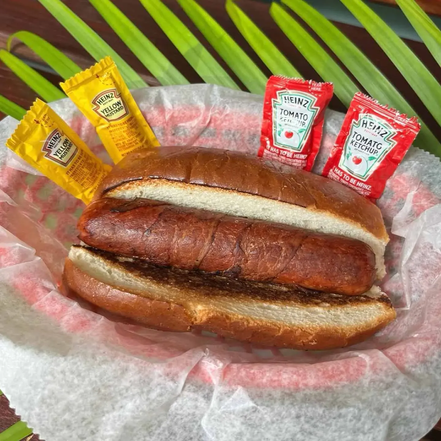 A grilled sausage in a bun with ketchup and mustard packets on paper in a basket, on a green leaf background.