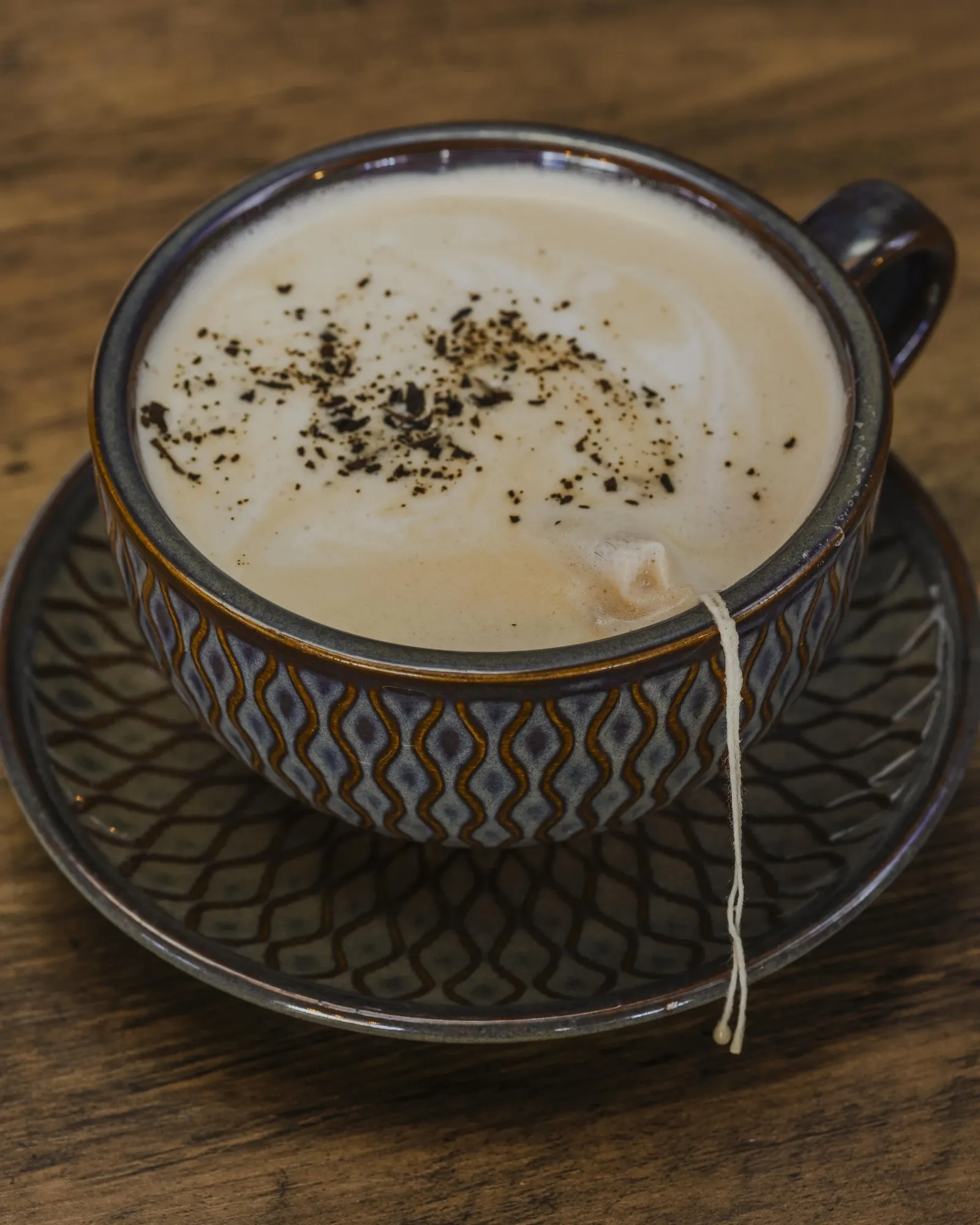 A cup of hot tea with milk and topped with cinnamon, a tea bag string can be seen hanging out of the top, the cup is sitting on a saucer and on a wood table.