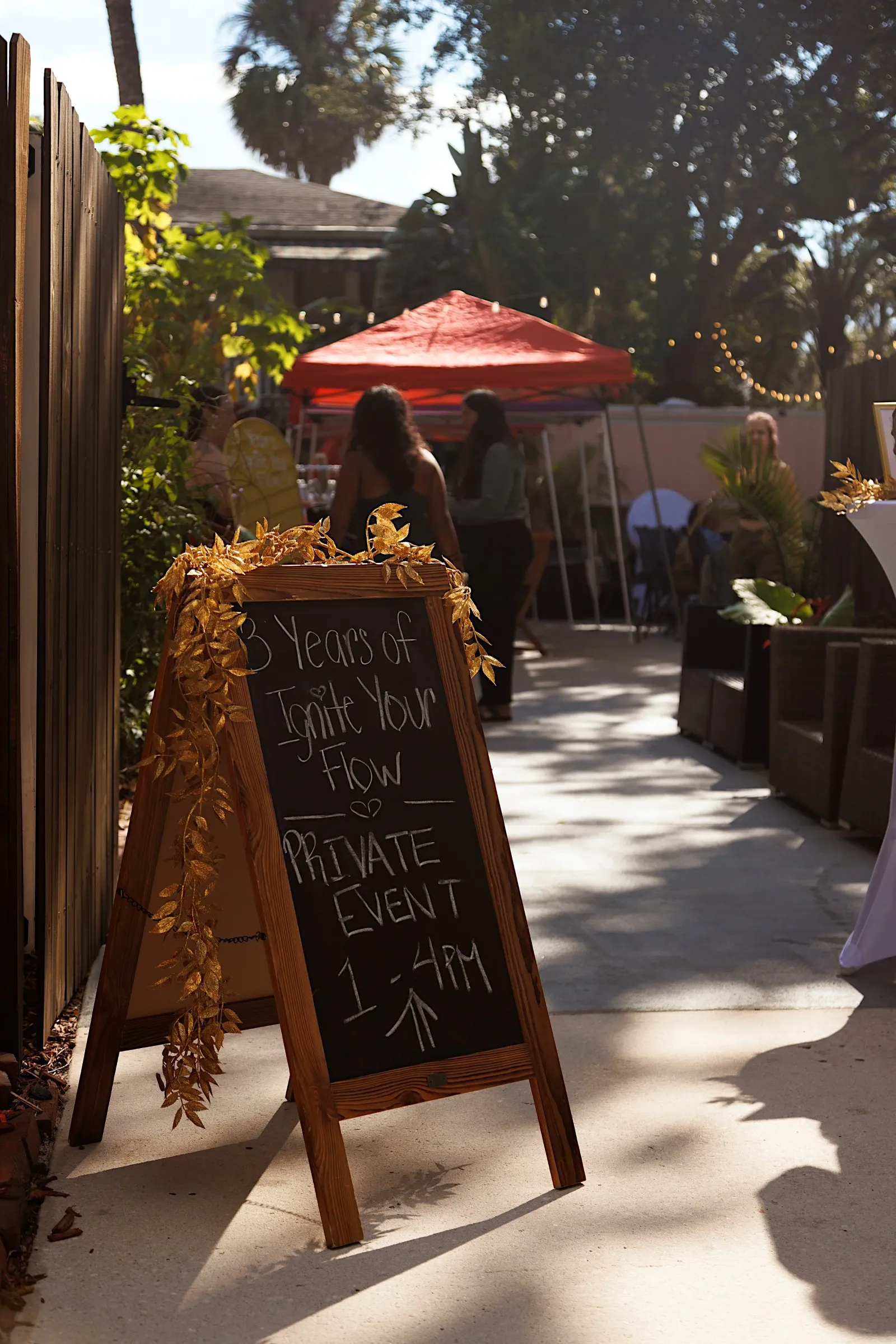 Outdoor private event sign with gold leaves decoration, advertising an event celebrating 3 years of something, with the time from 1 to 4 PM. In the background, there are people socializing under a red