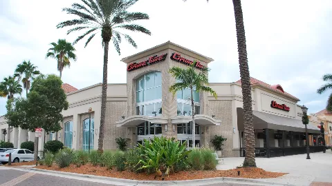 The outside of GameTime Fort Myers: a tan stucco building with a tall glass entrance, the red GameTime sign above it, palm trees and a planted bed in front