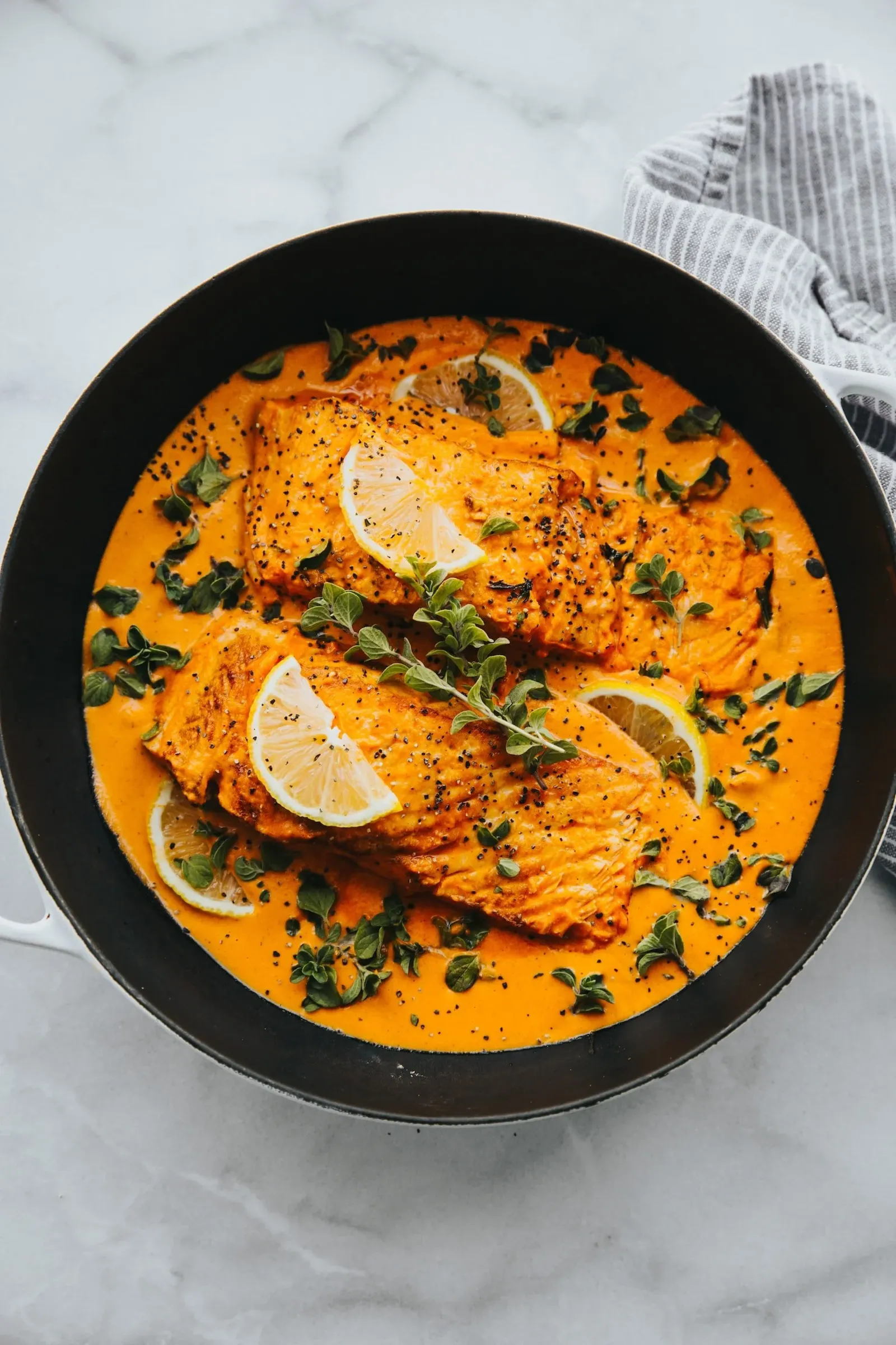Salmon fillets in a creamy orange sauce garnished with lemon slices and fresh herbs in a black skillet.