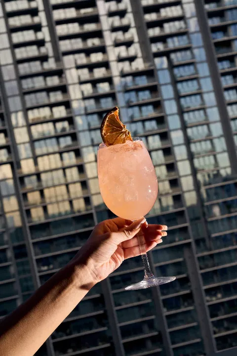 Pink cocktail in a stem glass with an orange slice garnish, held up against a blurred high-rise building backdrop.