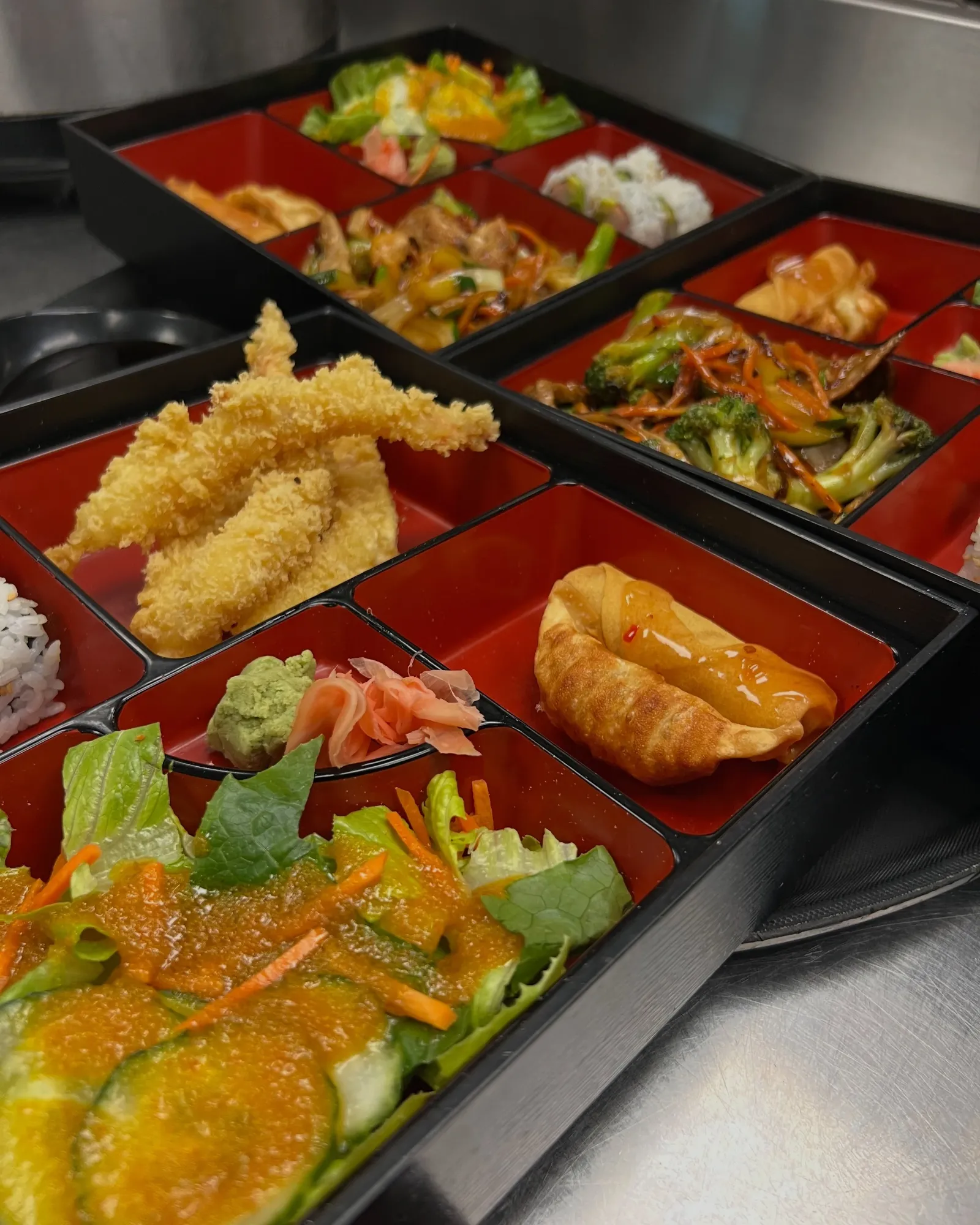 A Japanese bento box with various compartments containing salads, rice, tempura, gyoza, and vegetables.