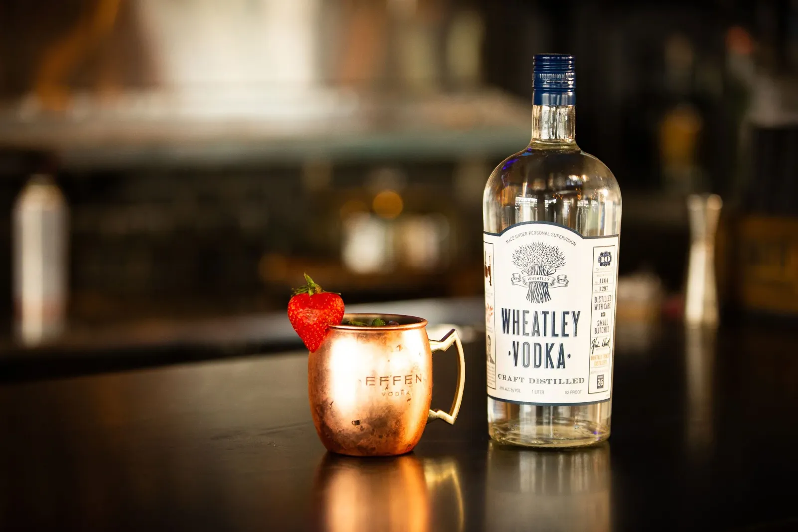 A bottle of Wheatley Vodka and a copper mug with a strawberry and mint on top, placed on a dark bar counter.