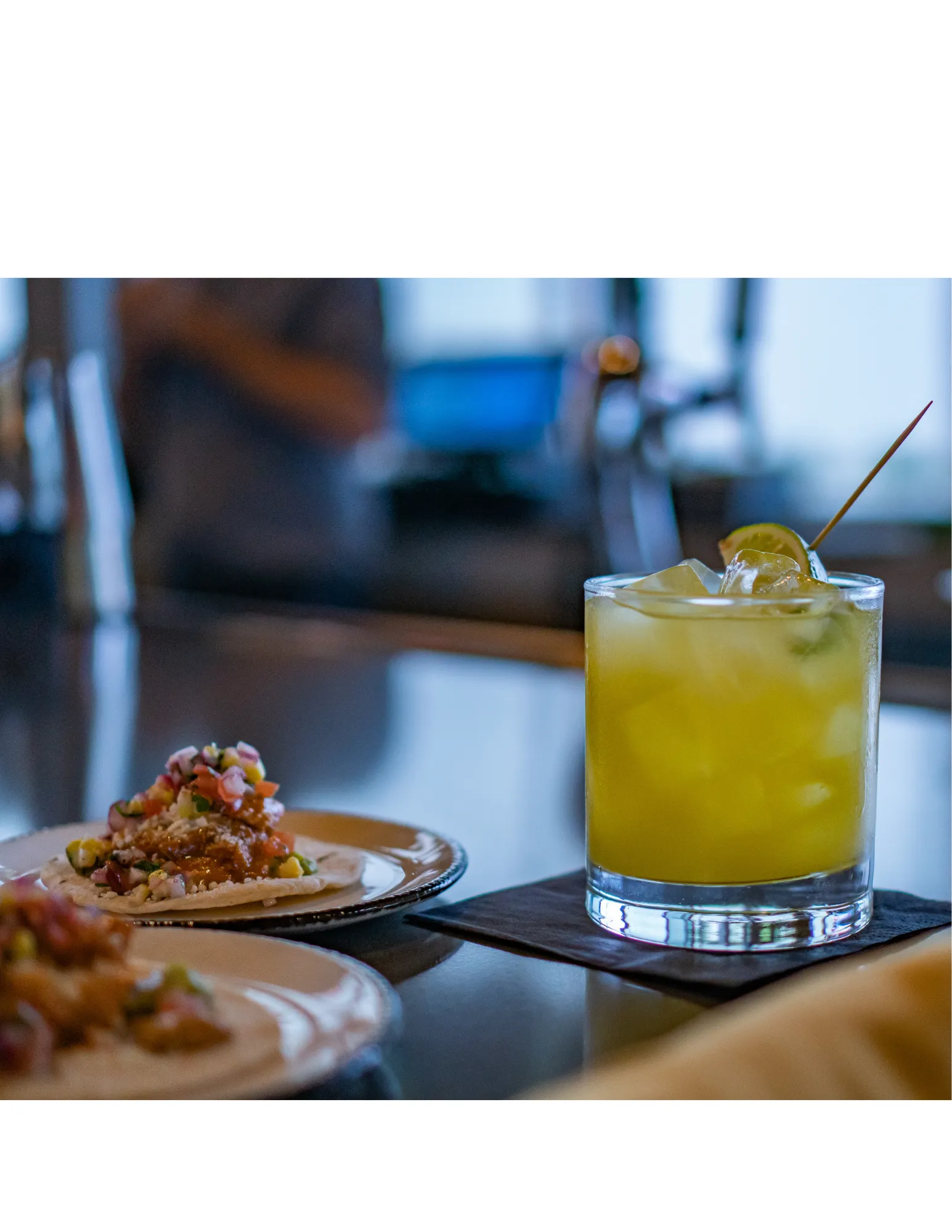 Close-up of a yellow cocktail with ice and lemon slices in a glass, with a plate of tacos topped with diced tomatoes, onions, and cilantro in the blurry background.