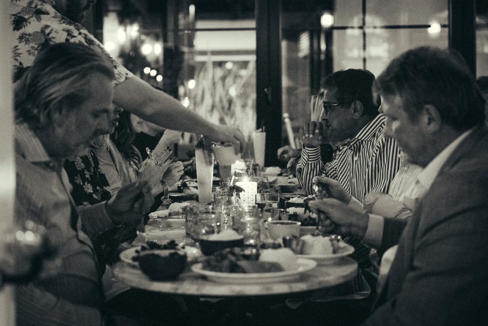 People sitting at a dinner table in a restaurant, engaged in conversation and eating, with glasses and candles on the table, in a dimly lit ambiance.