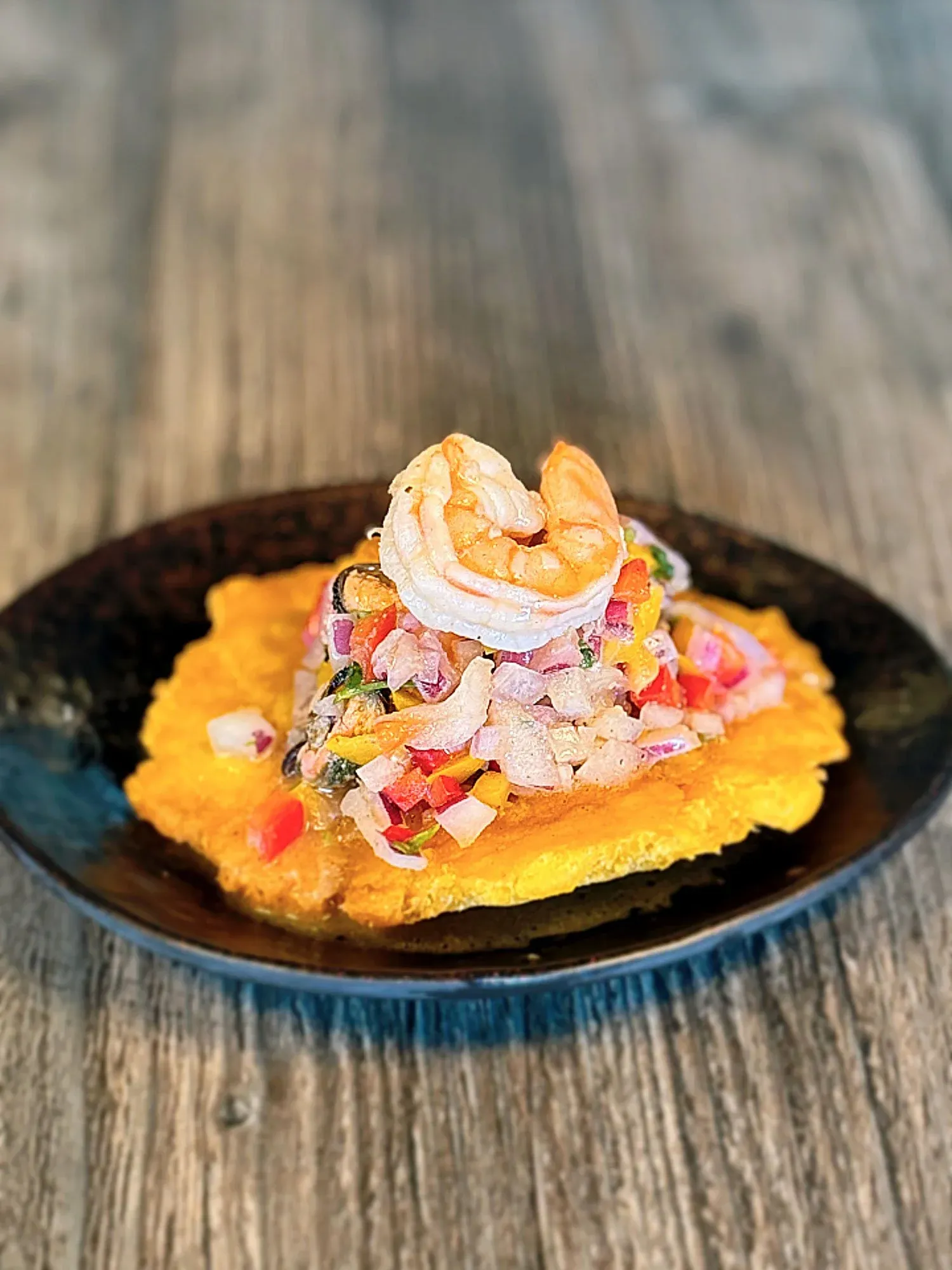 Shrimp on a bed of colorful diced vegetables served on a large plantain chip on a black plate.
