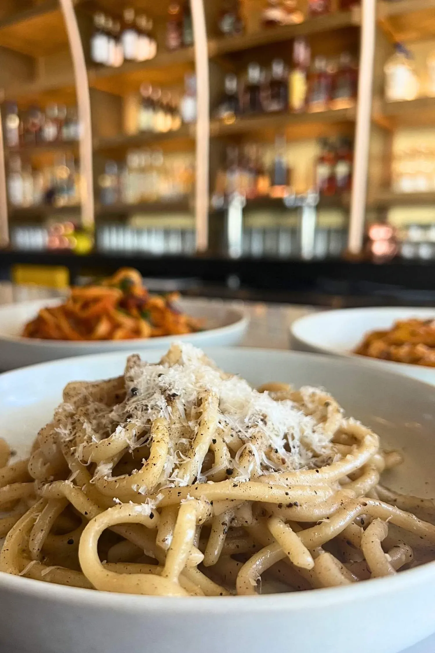 Spaghetti pasta topped with grated cheese and black pepper in a white bowl, with a blurred background of a kitchen shelving filled with jars.