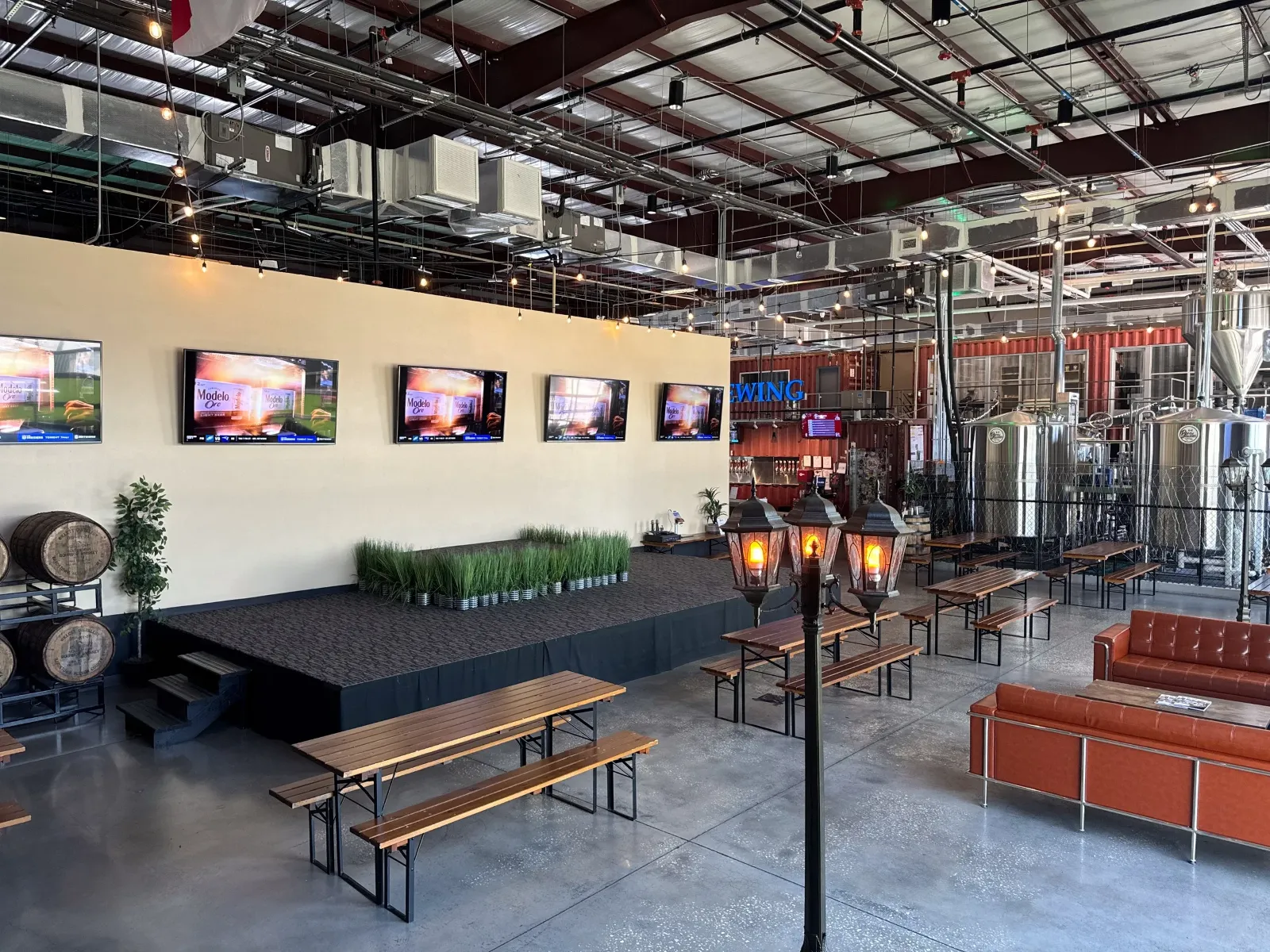 An interior view of a brewery or brewery taproom with a small stage, multiple screens, tables and benches, a cozy sofa, a street lamp-style light fixture, and brewing tanks.