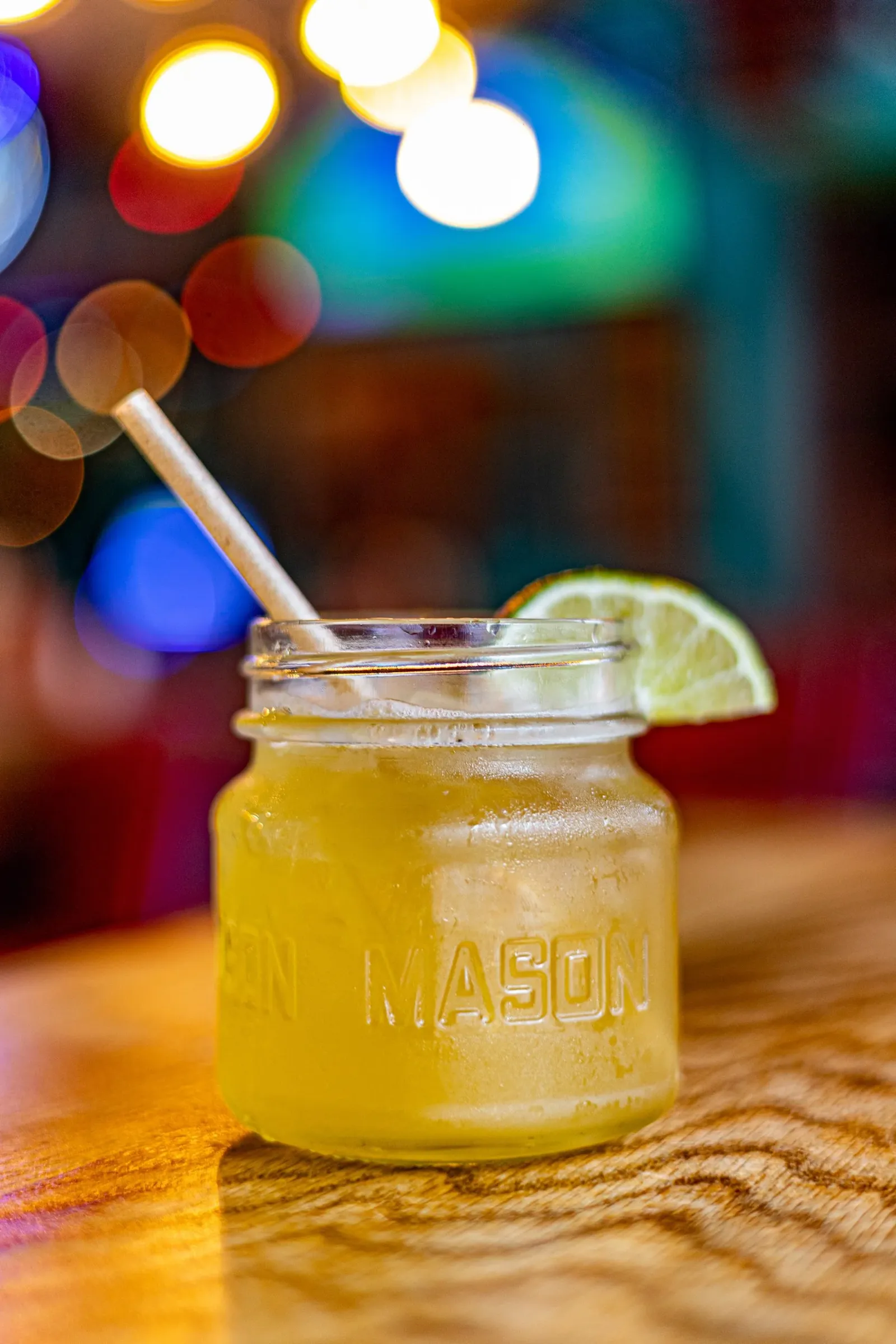 A mason jar filled with a yellow citrus drink, garnished with a lemon wedge, and a straw, on a wooden surface with colorful, blurry lights in the background.