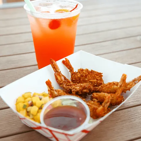 A tray of fried shrimp with dipping sauce, corn salad, and a large drink with ice and cherries on a wooden table.