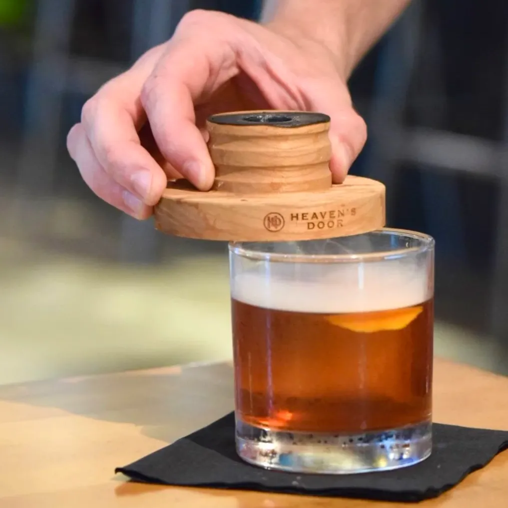 A hand holding a wooden object labeled 'Heaven's Door' over a glass of amber-colored liquid with a lemon slice inside, on a black napkin.