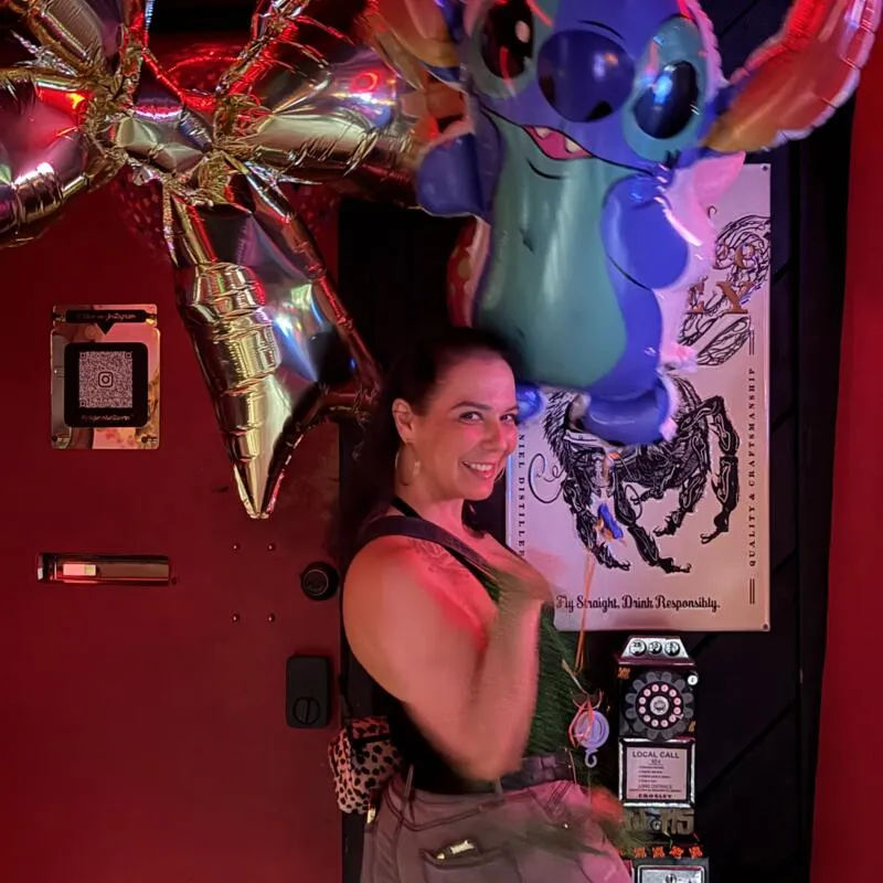 A person smiling in front of a festive backdrop decorated with balloons.