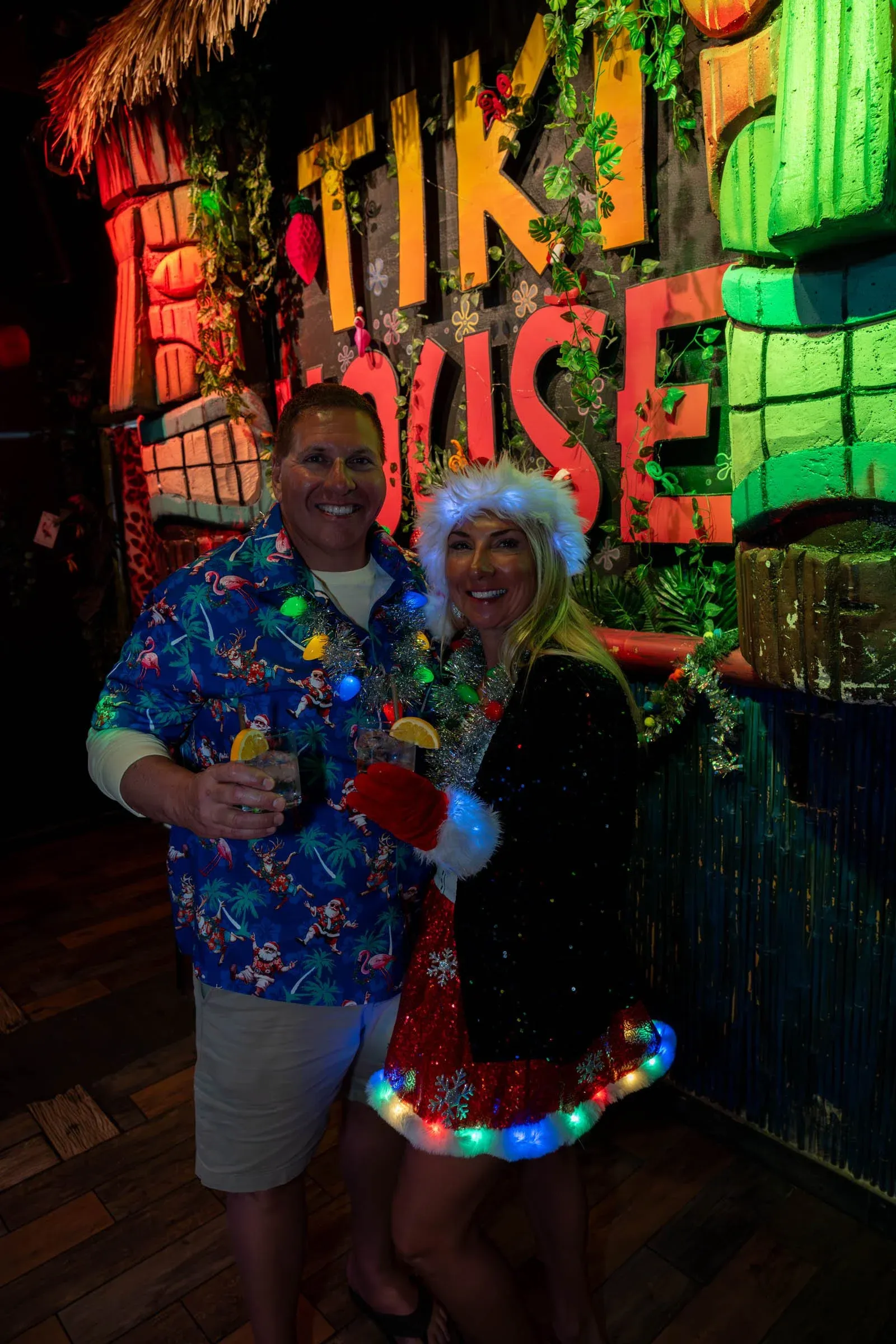 Two people celebrating Christmas at a festive party, wearing holiday attire and accessories, standing in front of a colorful sign that says TIKI, decorated with plants and holiday decorations.