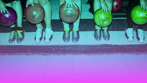 Children sitting on a bench at a bowling alley, holding colorful bowling balls, wearing bowling shoes, with the lane in front of them.