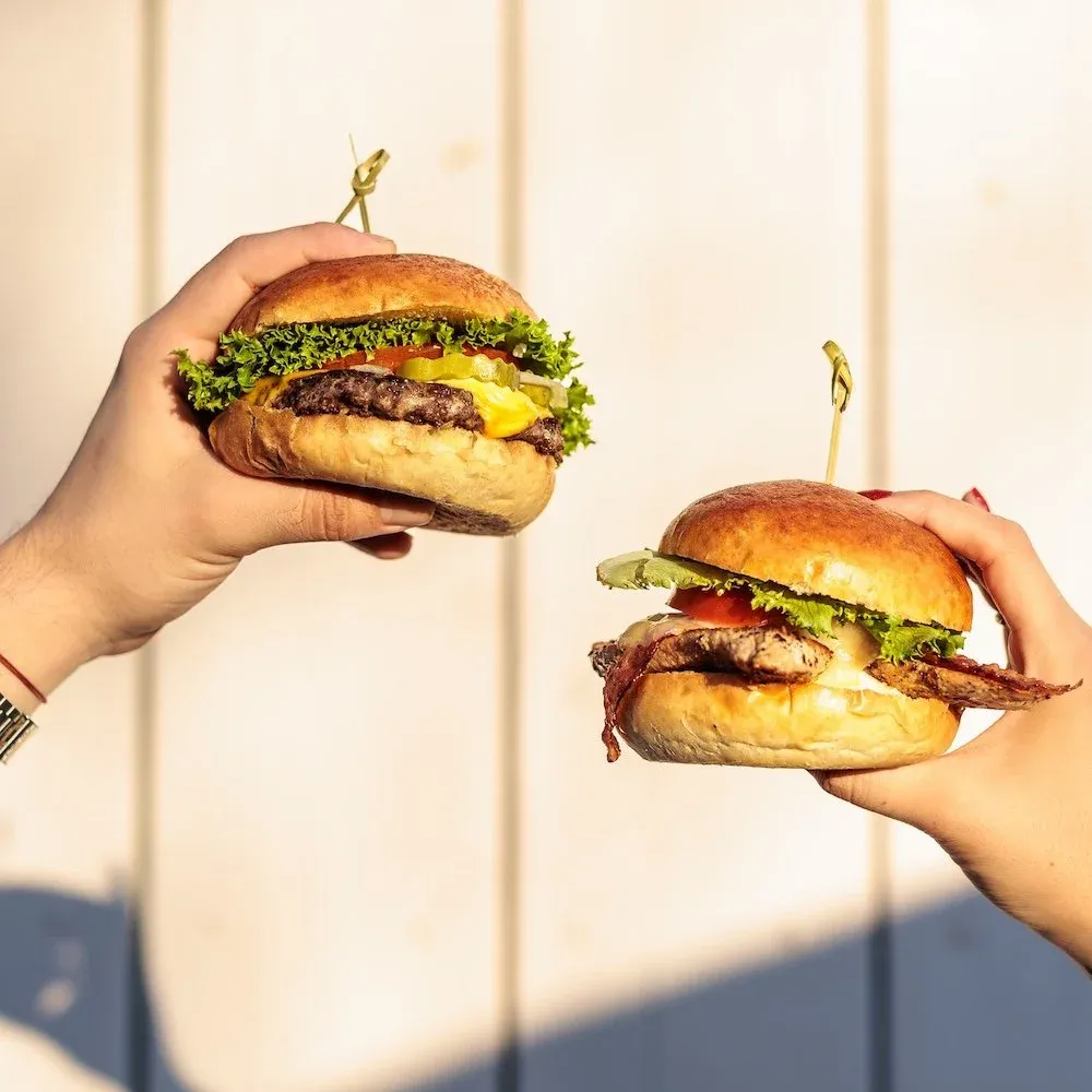 Two hands holding burgers with lettuce, tomato, pickle, cheese, and beef patties in buns, set against a light wooden background.