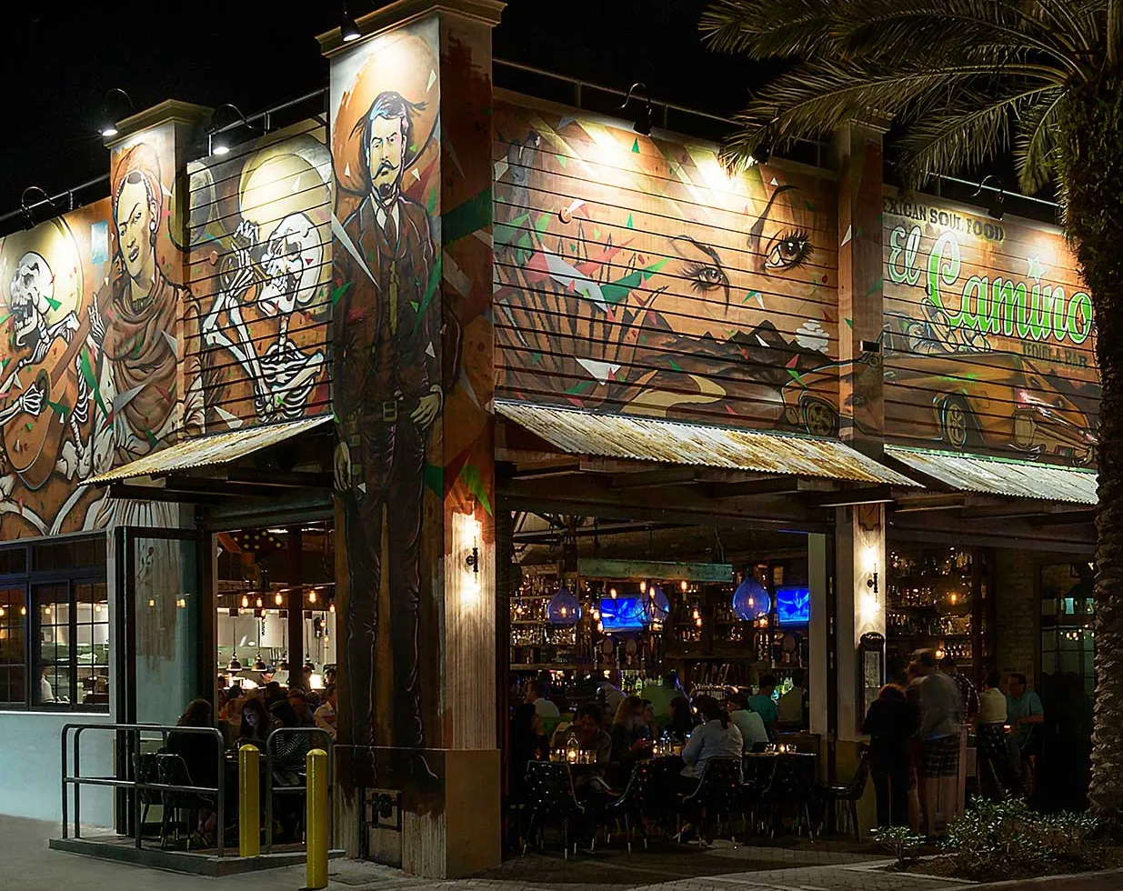 El Camino Delray exterior at night with colorful mural artwork of skeleton musicians and a man in a suit, corrugated metal awnings, and diners seated at outdoor tables