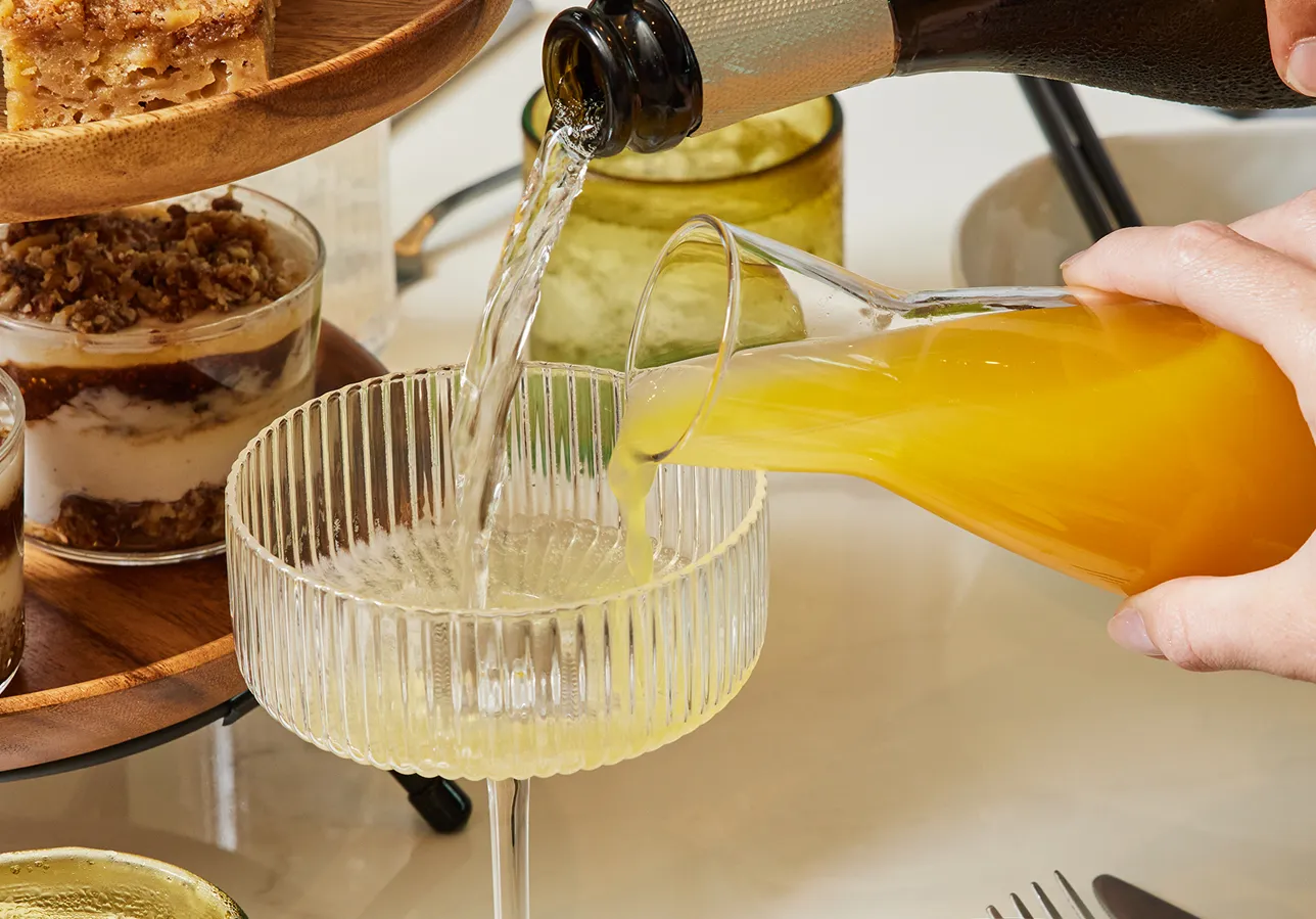 A person pours orange juice and sparkling wine into a glass. Dessert cups and baked goods are displayed on a tiered stand.