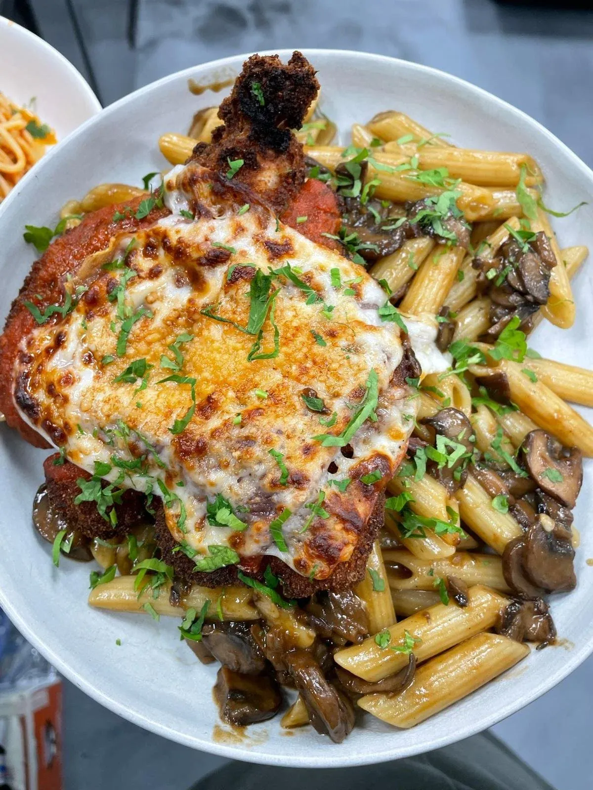 Baked chicken Parmesan with melted cheese, served with pasta and mushroom sauce, topped with chopped parsley.